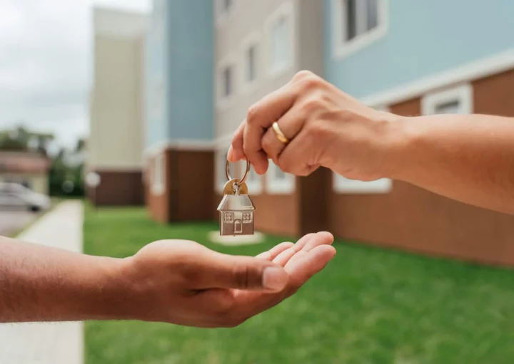 Imagem de uma pessoa com um anel de ouro entrega um chaveiro em forma de casa para outra em frente a um prédio de apartamentos e grama verde