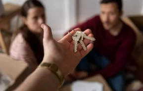 Imagem da mão de uma pessoa entregando uma chave em primeiro plano e um casal de jovens em segundo plano para ilustrar matéria sobre usufruto de imóvel