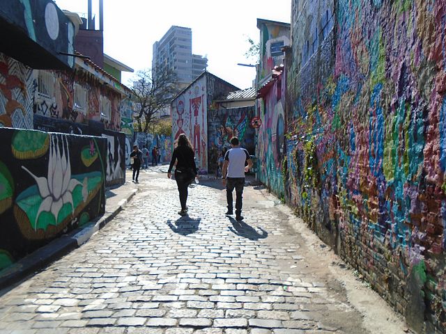 Duas pessoas caminham por uma rua de paralelepípedos ladeada por murais coloridos de grafite em SP em ambos os lados, sob luz solar intensa, com edifícios visíveis ao fundo.