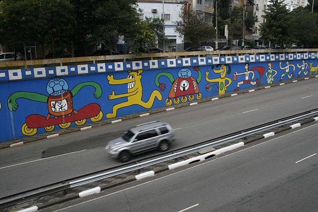 Um mural colorido inspirado no grafite em SP cobre um muro da cidade, mostrando animais de desenho animado em skates e carrinhos. Um SUV prateado passa pela rua, com árvores e prédios ao fundo.