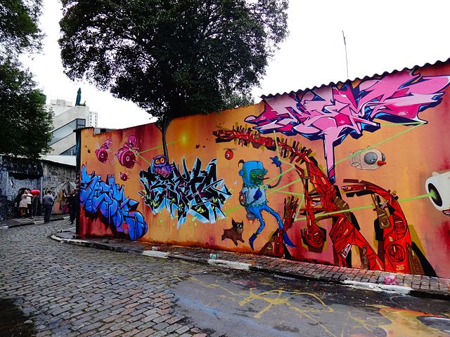 Um mural de rua colorido em uma parede ao longo de um beco de paralelepípedos em grafite em SP apresenta grafite abstrato, personagens de desenho animado, um gato e elementos robóticos em tons vibrantes de rosa, azul, laranja e vermelho sob uma árvore.