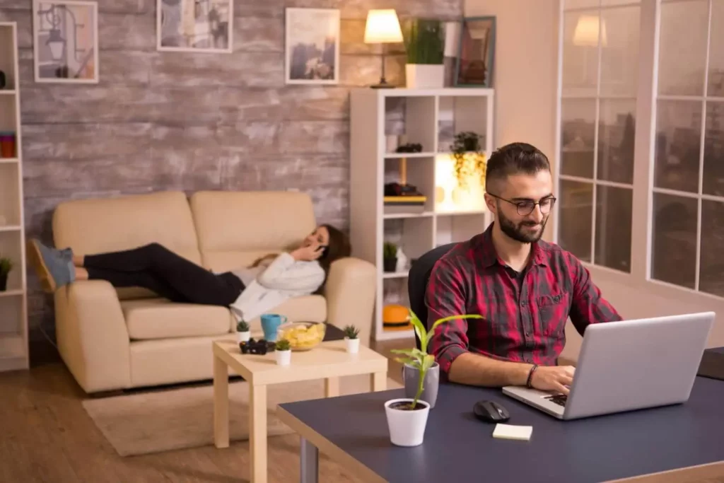 Imagem de um homem sentado em uma mesa trabalhando em um notebook enquanto uma mulher está deitada em um sofá da sala logo atrás falando ao telefone para ilustrar matéria sobre as cidades mais baratas do Brasil para se viver