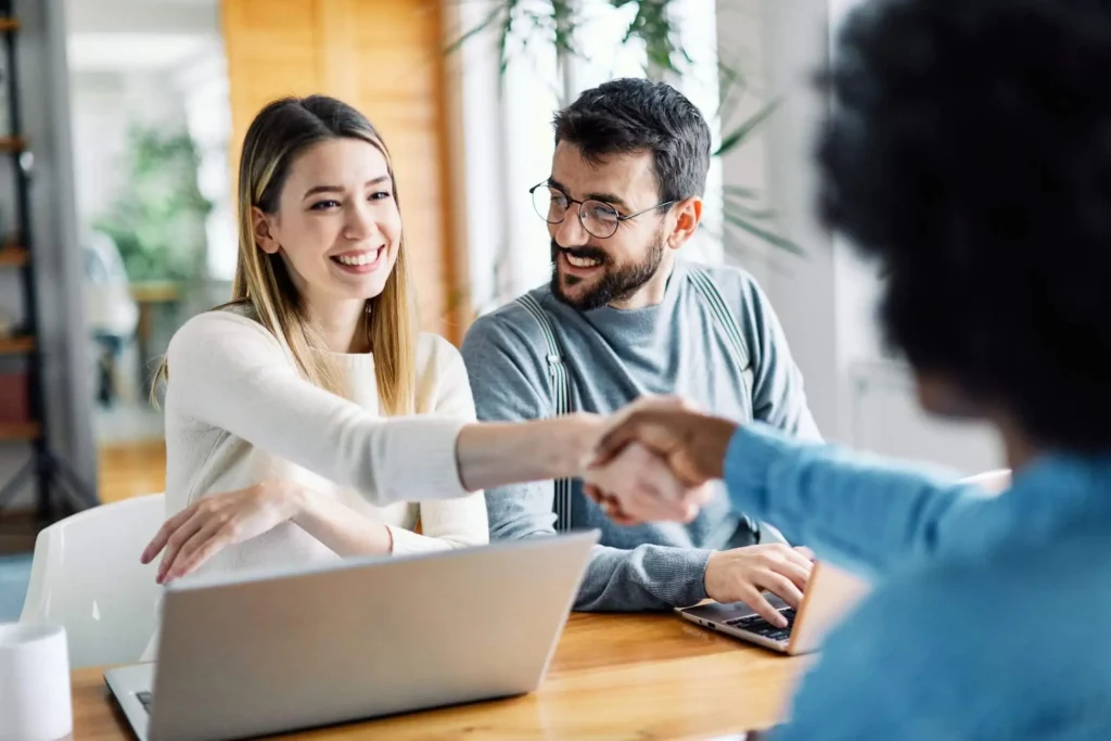 Imagem de duas pessoas sentadas em uma mesa com laptops, sorrindo enquanto uma mulher aperta a mão de uma terceira pessoa que está de costas para a câmera, sugerindo uma reunião amigável sobre como financiar um imóvel ou um acordo comercial bem-sucedido