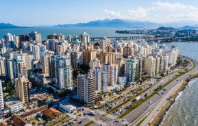 Foto que ilustra matéria sobre custo de vida em Florianópolis mostra uma visão aérea de vários prédios da cidade à beira-mar em um dia ensolarado, com uma ponte ao fundo.