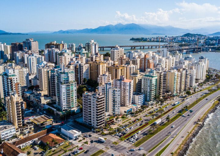 Foto que ilustra matéria sobre custo de vida em Florianópolis mostra uma visão aérea de vários prédios da cidade à beira-mar em um dia ensolarado, com uma ponte ao fundo.