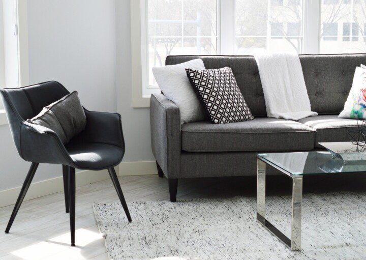 Imagem que ilustra matéria sobre decoração modernista mostra o detalhe de uma sala com uma poltrona preta, metade de um sofá cinza escuro com almofadas em cima e parte de uma mesa de centro de tampo de vidro sobre um tapete