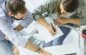 Duas pessoas estão sentadas a uma mesa de vidro, trabalhando juntas. Uma escreve em um papel, enquanto a outra aponta para a tela de um laptop. Uma calculadora, papéis e uma caneca de café vermelha também estão sobre a mesa. A imagem é de cima.