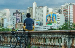 Foto que ilustra matéria sobre grafite em BH mostra o mirante da Rua Sapucaí, localizada no Centro da Cidade, de onde se vê ao fundo diversos prédios, alguns deles com artes coloridas pintadas em suas fachadas ou laterais. Sobre a mureta, com uma bicicleta encostada, um homem sentado de costas e de pernas cruzadas observa as obras de arte.