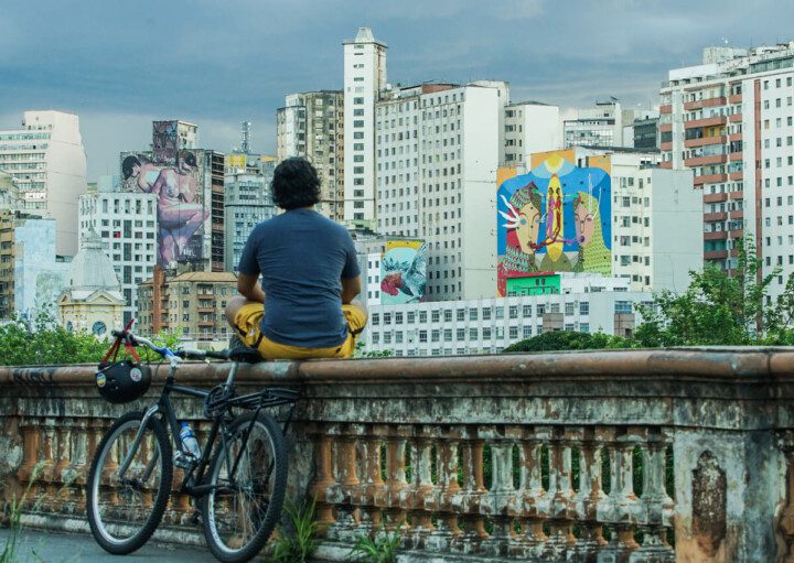 Foto que ilustra matéria sobre grafite em BH mostra o mirante da Rua Sapucaí, localizada no Centro da Cidade, de onde se vê ao fundo diversos prédios, alguns deles com artes coloridas pintadas em suas fachadas ou laterais. Sobre a mureta, com uma bicicleta encostada, um homem sentado de costas e de pernas cruzadas observa as obras de arte.