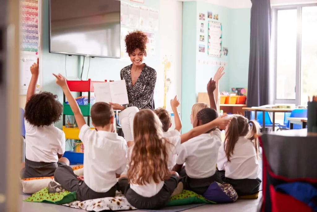 Imagem de uma professora apresentando um livro para crianças que estão sentadas em almofadas no chão com as mãos para cima em uma sala de aula