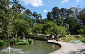 foto do parque celso daniel, na cidade de santo andré. Mulher e homem caminham perto de lago, com fontes, cercado pela mata