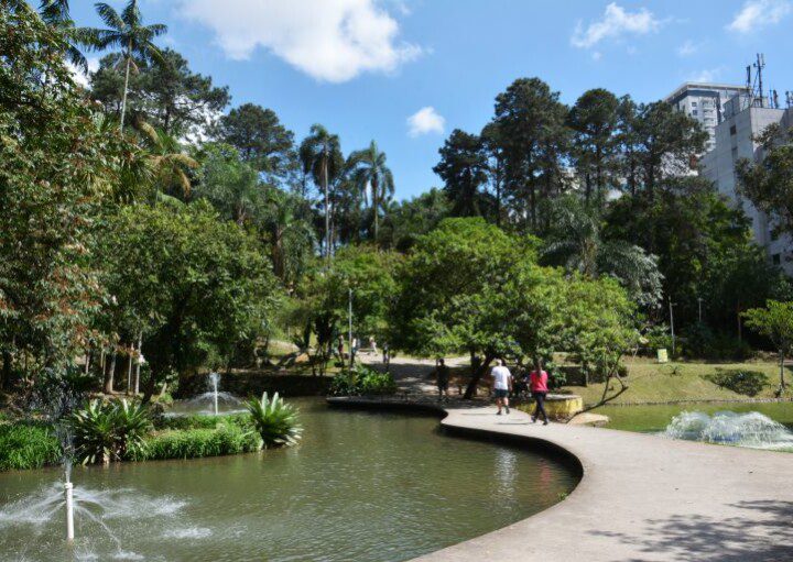 foto do parque celso daniel, na cidade de santo andré. Mulher e homem caminham perto de lago, com fontes, cercado pela mata