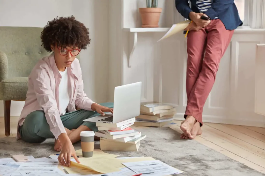 Imagem de uma mulher sentada em cima de um tapete que está no chão de uma sala junto a papéis, livros, um notebook e um copo de café descartável para ilustrar matéria sobre qual a cidade mais barata para se viver no Brasil