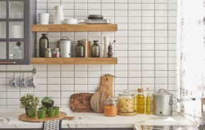 Foto que ilustra matéria sobre revestimento para cozinha mostra uma parede de azulejos brancos pequenos com duas prateleiras de madeira repletas de potes de vidro, cerâmica e metal. Além de uma bancada de mármore branco com vasos pequenos de plantas, tábuas de corte, potes com macarrão e grãos e vidros de azeite.