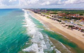 Foto que ilustra matéria sobre praias de Fortaleza mostra, de um ângulo do alto, boa parte da extensão da Praia do Futuro, com o mar aparecendo ao lado esquerdo da tela e a faixa de terra, com areia, algumas casas e vegetação, ao lado direito. (Foto: Jade Queiroz | MTUR)