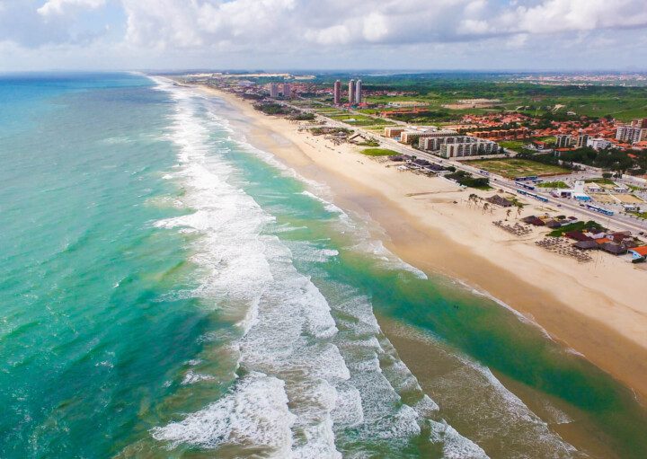 Foto que ilustra matéria sobre praias de Fortaleza mostra, de um ângulo do alto, boa parte da extensão da Praia do Futuro, com o mar aparecendo ao lado esquerdo da tela e a faixa de terra, com areia, algumas casas e vegetação, ao lado direito. (Foto: Jade Queiroz | MTUR)