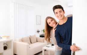 Foto que ilustra matéria sobre chá de casa nova mostra um casal sorridente abrindo a porta de uma com uma sala ao fundo, onde aparece um sofá, uma mesa de centro e uma janela com cortina na frente.