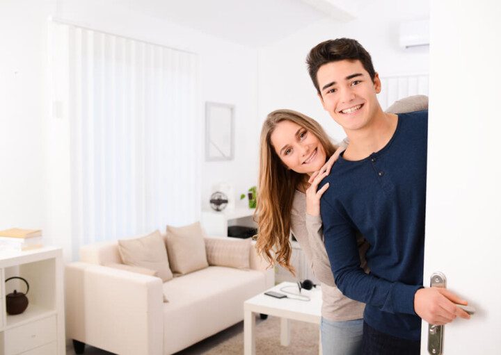 Foto que ilustra matéria sobre chá de casa nova mostra um casal sorridente abrindo a porta de uma com uma sala ao fundo, onde aparece um sofá, uma mesa de centro e uma janela com cortina na frente.