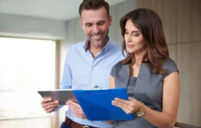 Imagem de dois profissionais de negócios estão em um ambiente interno, sorrindo e analisando documentos juntos, para ilustrar mtéria sobre consultoria imobiliária. O homem segura um tablet e a mulher segura uma prancheta azul em um ambiente de escritório moderno.