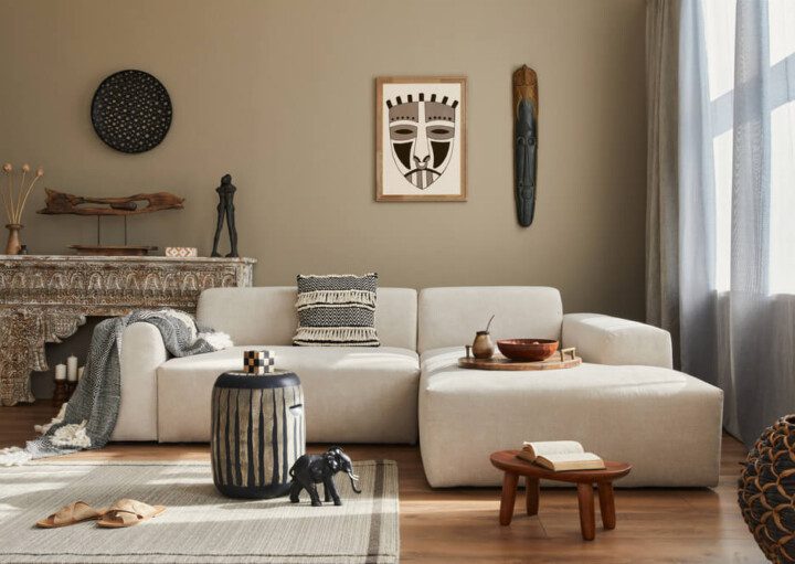 Foto que ilustra matéria sobre cores de parede para sala mostra uma sala com uma parede bege ao fundo, um sofá branco à frente e diversos objetos de decoração, tanto na parede quanto em uma estante entre a parede e o sofá.