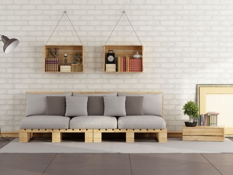 Sofá de palete com estofado, encostado em uma parede de tijolinhos, com dois nichos pendurados nela