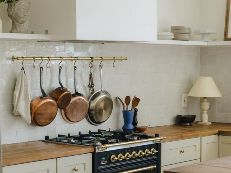 A foto mostra uma bancada, com um fogão ao centro. Há panelas penduradas na parede e armários de teto. Tem também outros elementos decorativos como uma abajour, utensílios de cozinha e louças bonitas. 