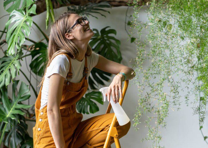 Foto que ilustra matéria sobre plantas pendentes mostra uma mulher loira, de cabelos lisos, magra e de óculos. Sorridente, ela está no alto de uma escada, com um borrifador de água para regar plantas de diversos tipos de pendem do alto na imagem.