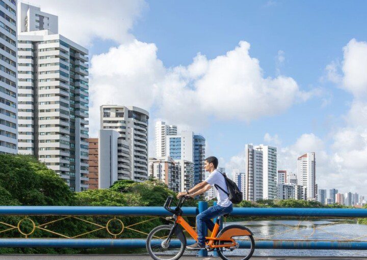 homem andando de bicicleta às margens de um rio, com prédios na lateral esquerda da foto em Recife