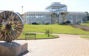 Foto que ilustra matéria sobre parques em Sorocaba mostra o Jardim Botânico Irmãos Vilas Bôas, com uma escultura redonda feita em metal à esquerda da imagem, em primeiro plano, e mais ao fundo uma estrutura de vidro conhecida como Palacete de Cristal