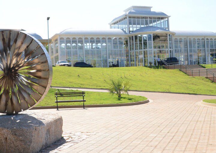 Foto que ilustra matéria sobre parques em Sorocaba mostra o Jardim Botânico Irmãos Vilas Bôas, com uma escultura redonda feita em metal à esquerda da imagem, em primeiro plano, e mais ao fundo uma estrutura de vidro conhecida como Palacete de Cristal