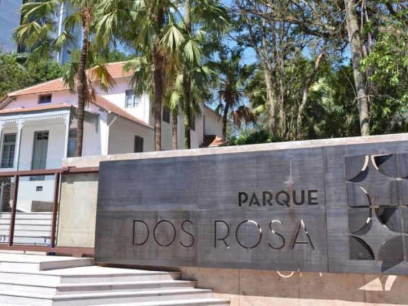 Placa de concreto da entrada do Parque do Rosa. Ao fundo um casarão branco e uma árvore.