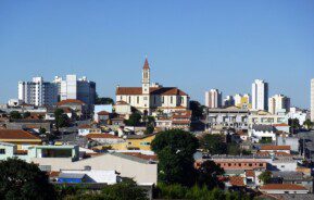 Foto que ilustra matéria sobre a Zona Leste de SP mostra uma panorâmica do bairro de Vila Matilde, com uma igreja ao fundo, no centro da tela, e um céu azul.