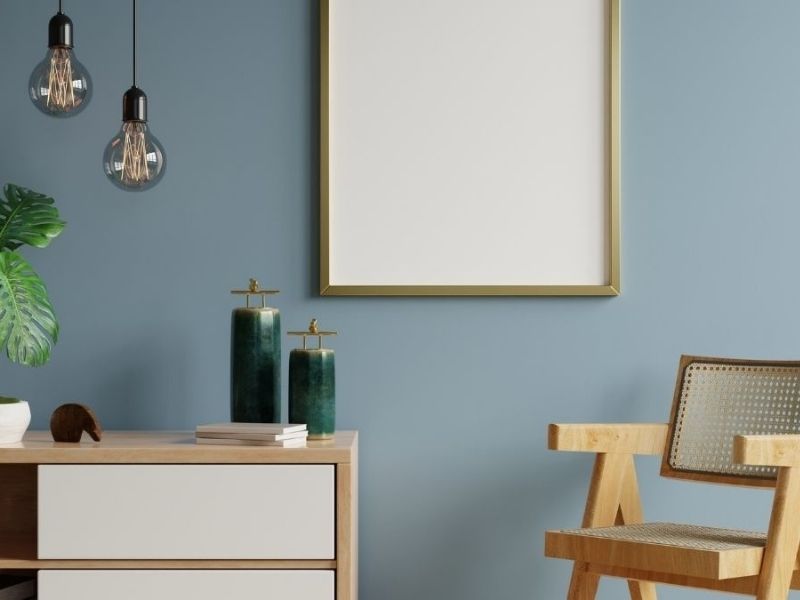 parede azul, espelho pendurado nela. Abaixo uma cômoda com itens decorativos e, ao lado da cômoda, uma cadeira