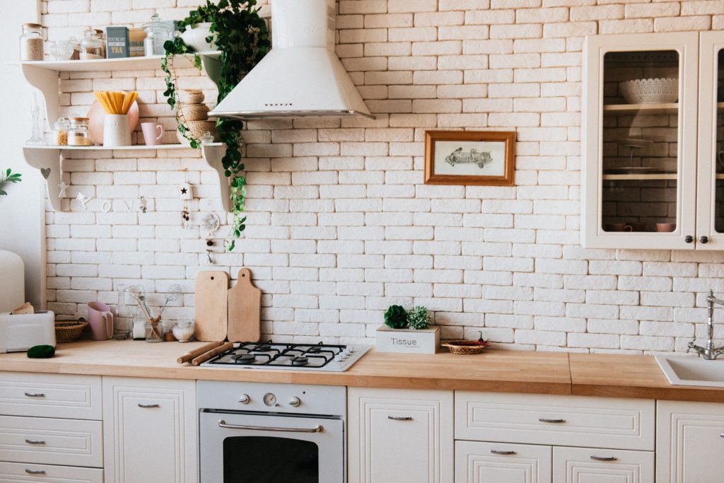 Cozinha toda na cor branca, com parede de tijolinhos, fogão, exaustor, cabines com porta de vidro e plantas de decoração