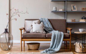 sala de estar decorada no estilo japandi. Sofá de dois lugares, cinza, com manta azul caida no chão. Ao lado, vaso com flores e mesinha redonda. Atrás, prateleira com objetos decorativos