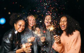 Foto que ilustra matéria sobre resoluções de ano novo mostra quatro jovens mulheres em uma festa de Réveillon, sorridentes e com velas que soltam faíscas nas mãos, enquanto confetes coloridos voam pelos ares diante delas.