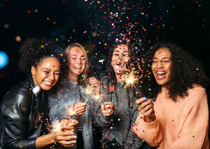 Foto que ilustra matéria sobre resoluções de ano novo mostra quatro jovens mulheres em uma festa de Réveillon, sorridentes e com velas que soltam faíscas nas mãos, enquanto confetes coloridos voam pelos ares diante delas.