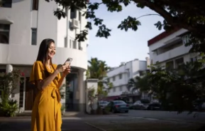 Imagem de uma mulher parada em uma rua residencial em frente a alguns prédios mexendo no celular para ilustrar matéria sobre a Zona Norte do Rio de Janeiro