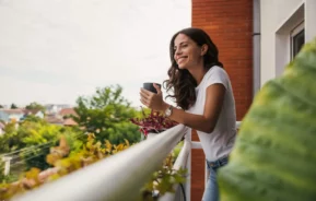 Imagem de uma mulher com longos cabelos escuros, vestindo uma camiseta branca e calça jeans, em uma varanda segurando uma caneca e sorrindo. A vegetação e as árvores preenchem o fundo em um dia claro