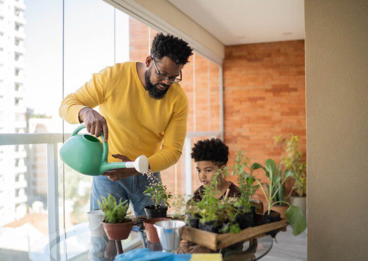 Foto que ilustra matéria sobre Como fazer horta em casa mostra um homem negro, de camisa amarela comprida, usando um regador para molhar pequenos vasos de plantas com ervas ao lado de seu filho em uma sacada de apartamento