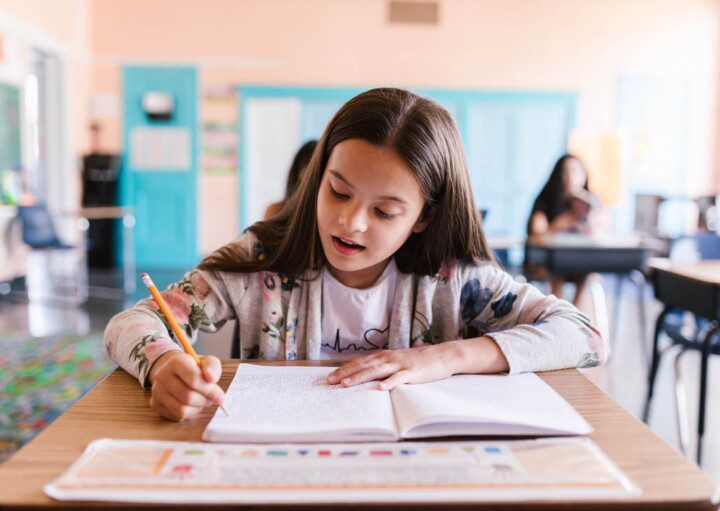 Foto que ilustra matéria sobre Escolas em Vitória (ES) mostra uma menina sentada com um caderno aberto à sua frente e escrevendo com um lápis amarelo em uma sala de aula.