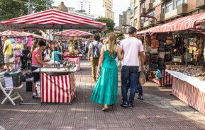 Foto que ilustra matéria sobre a Feira da Liberdade mostra um trecho da feira com barracas dos dois lados e pessoas passando pelo meio