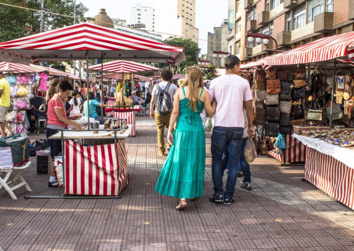 Foto que ilustra matéria sobre a Feira da Liberdade mostra um trecho da feira com barracas dos dois lados e pessoas passando pelo meio