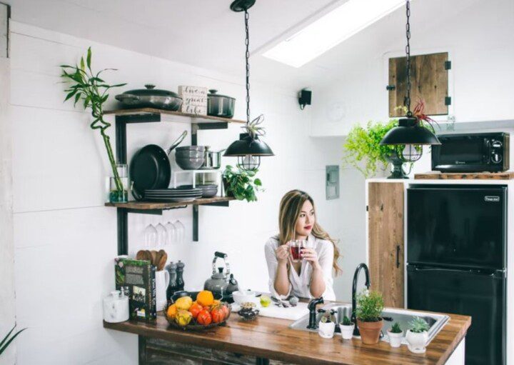 A foto mostra uma mulher sentada à mesa de uma cozinha pequena e bem decorada. Na imagem há uma bancada com pia, frutas, plantas, livro, chaleira e temperos. Há também na imagem uma prateleira com utensílios de cozinha (pratos, panelas e outros), além de uma geladeira.