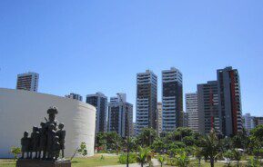 Foto que ilustra matéria sobre parques em Recife mostra uma panorâmica do Parque Dona Lindu, com uma escultura em primeiro plano, mais à esquerda, árvores à direita e grandes prédios residenciais ao fundo.