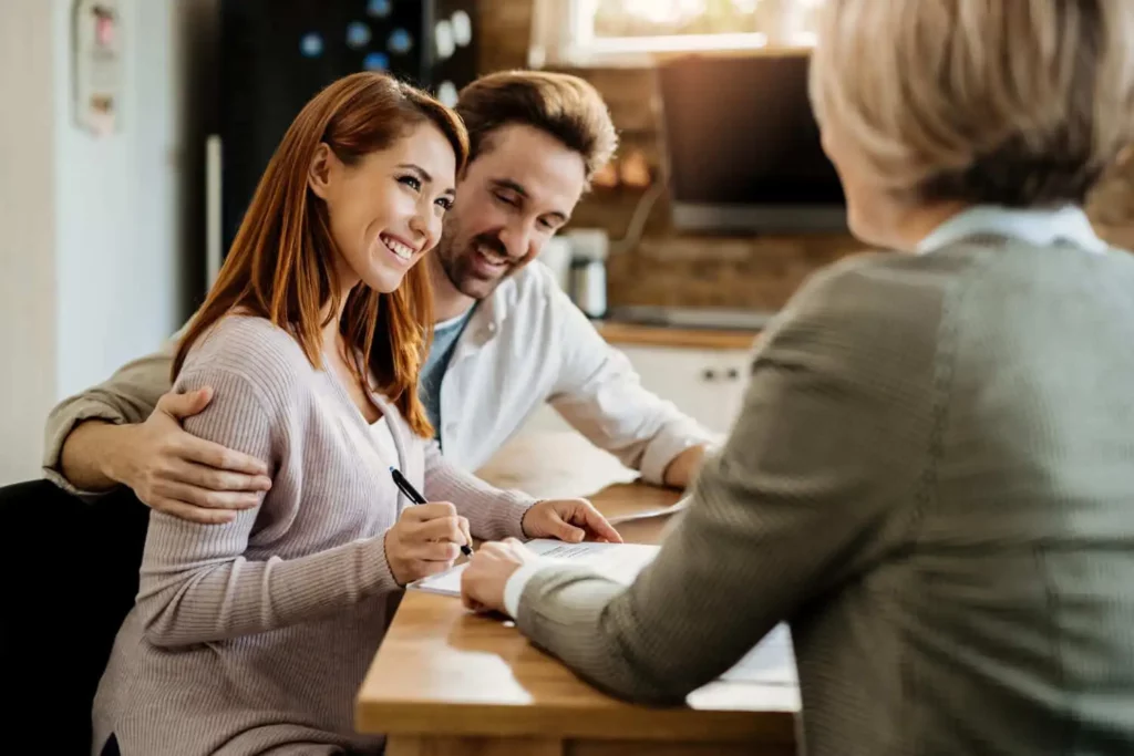 Imagem de um casal sorridente sentado em uma mesa, com a mulher assinando alguns papéis, enquanto uma pessoa em frente a eles, de costas para a câmera, supervisiona os documentos. O casal parece feliz e animado.