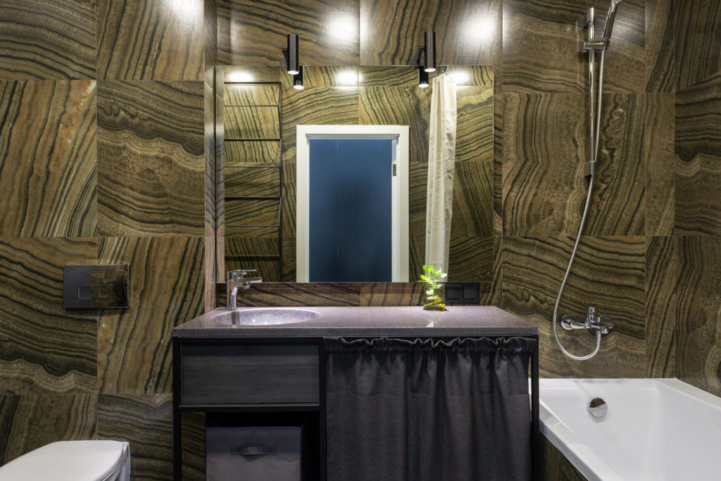 Foto de banheiro pequeno decorado com texturas na parede. Nelas foi utilizado um revestimento em tons dourados e pretos. Há também: bancada, pia, banheira, ducha, vaso sanitário e espelho.