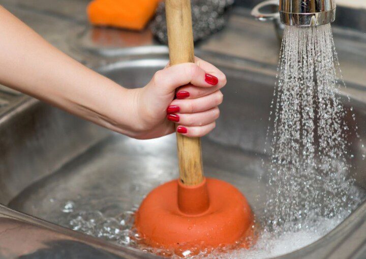 Mão com unhas pintadas de esmalte segurando um desentupidor dentro de uma pia de cozinha entupida, ensinando como desentupir pia de cozinha