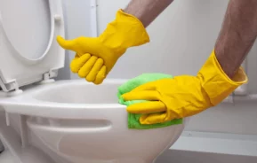 Imagem de um homem limpando um vaso sanitário com luvas amarelas e fazendo sinal de joinha para ilustrar matéria sobre como desentupir vaso sanitário