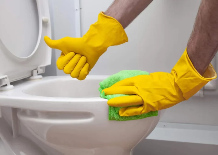 Imagem de um homem limpando um vaso sanitário com luvas amarelas e fazendo sinal de joinha para ilustrar matéria sobre como desentupir vaso sanitário